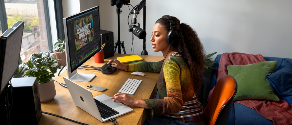 Una donna che lavora da casa seduta alla scrivania con un MacBook Pro collegato a due monitor esterni, che indossa un paio di cuffie e parla in un microfono