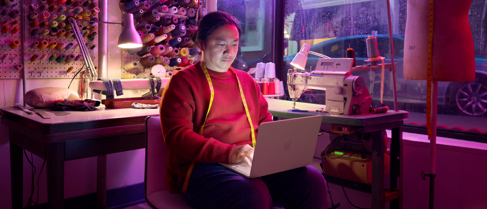 Una persona che lavora tenendo sulle ginocchia un MacBook Neo rosa pastello non collegato alla corrente, in un ambiente con una macchina da cucire e relativi materiali, tra cui filo e manichino