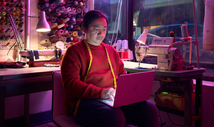 Una persona che lavora tenendo sulle ginocchia un MacBook Neo rosa pastello non collegato alla corrente, in un ambiente con una macchina da cucire e relativi materiali, tra cui filo e manichino