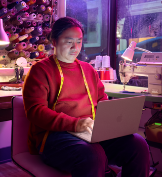 Una persona che lavora tenendo sulle ginocchia un MacBook Neo rosa pastello non collegato alla corrente, in un ambiente con una macchina da cucire e relativi materiali, tra cui filo e manichino